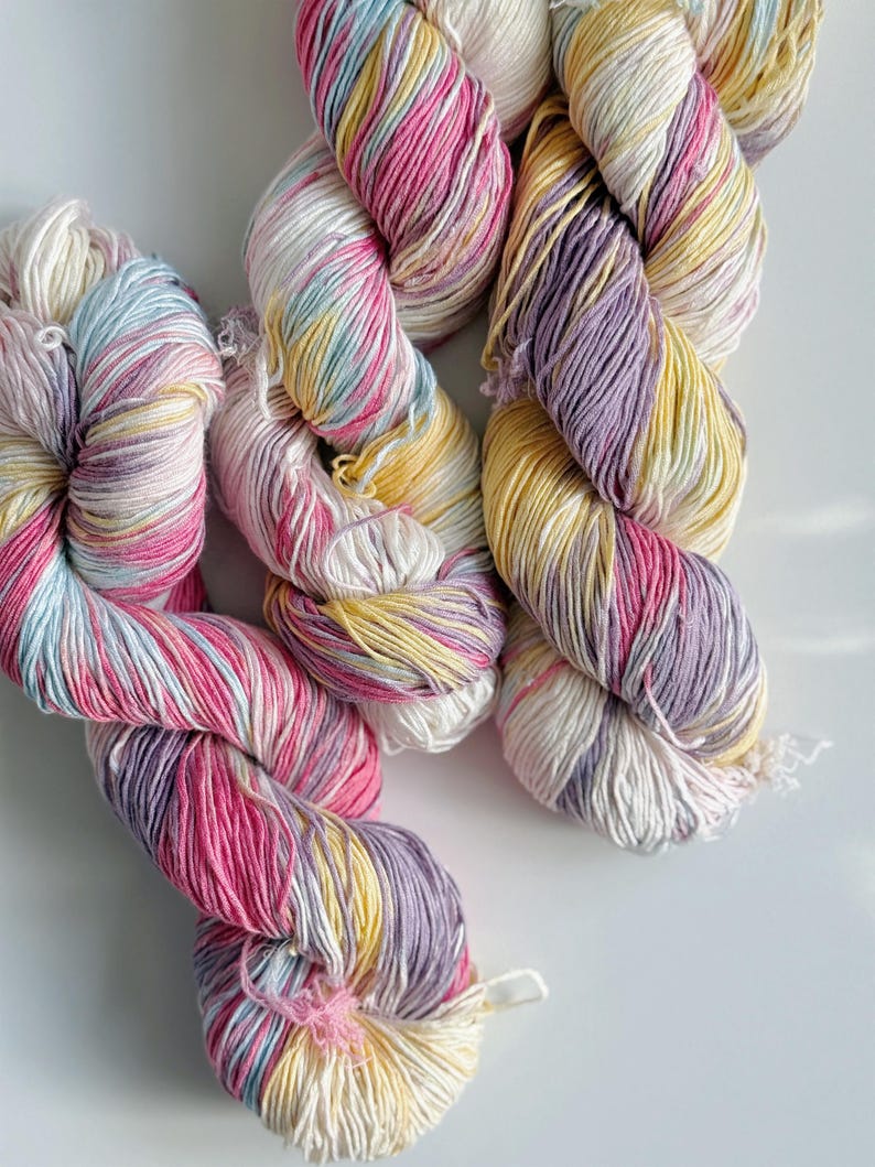 Puede incluir: Primer plano de varios ovillos de hilo colorido. El hilo es una mezcla de rosa, amarillo, morado y blanco. El hilo est&aacute; torcido y parece suave, ideal para proyectos de tejido o ganchillo.