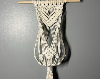May include: Cream-colored macrame plant hanger with a clear glass globe. The hanger is suspended from a wooden dowel and features intricate knotting patterns. A long tassel hangs from the bottom. Perfect for displaying small plants.