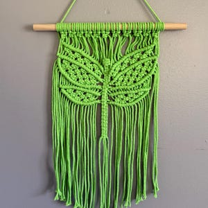 May include: A vibrant green macrame wall hanging featuring a dragonfly design. The handmade piece is suspended from a natural wooden dowel and includes long, fringed tassels. This decorative item adds a touch of bohemian style to any space.