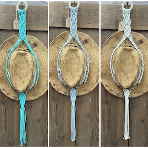 May include: Three macrame plant hangers with a woven design and a tassel at the bottom. The hangers are in different colors: turquoise, light gray, and white. The hangers are hanging from a wooden surface.