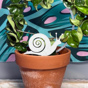 May include: A silver-colored snail-shaped plant trellis insert featuring a spiral shell design. The trellis is positioned inside a terracotta-colored clay pot containing a green Hoya plant with thick, waxy leaves wrapped around a circular support.