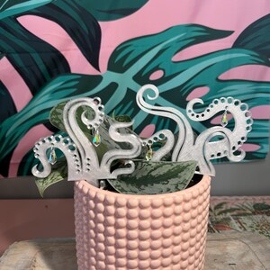 May include: A light pink, textured ceramic pot holds a green plant with silver octopus tentacle plant stakes. The stakes have dangling iridescent crystals. The background features a pink and green tropical leaf pattern.