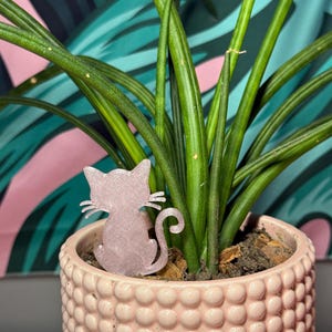May include: A pink cat-shaped plant stake is placed in a pink, textured ceramic pot with a green plant. The pot has a rounded, dotted pattern. The background features a pink and teal patterned fabric.