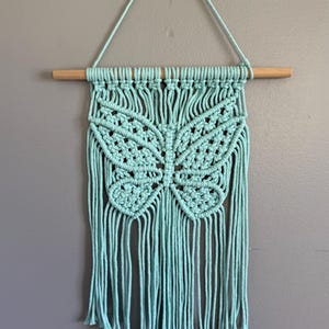 May include: A turquoise macrame wall hanging featuring a butterfly design. The butterfly is centered, with detailed knotting. The piece is suspended from a natural wooden dowel and hanging cord, adding a touch of bohemian decor.