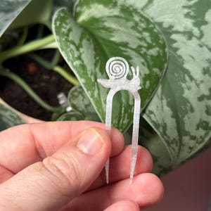 May include: A silver-colored hair fork with a snail design. The fork has a U-shaped base with two prongs and a snail figure at the top. The background features green and silver plant leaves.