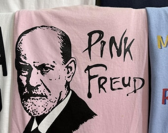 May include: Pink t-shirt featuring a black and white portrait of Sigmund Freud with the words "Pink Freud" in black lettering. The shirt is folded and surrounded by other t-shirts with various designs.