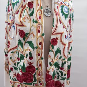 May include: Cream-colored embroidered shawl with a floral design featuring red pomegranates, green leaves, and gold and red accents. A silver pendant with a tree of life design hangs from a delicate chain.