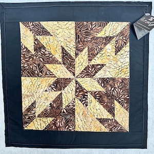 May include: A square quilt with a central starburst pattern in shades of brown and yellow, framed by a black border. The starburst design features leaf-like patterns. The quilt is likely handmade and suitable for home decor.