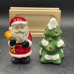 May include: A ceramic Christmas salt and pepper shaker set. One shaker is Santa Claus in a red suit with a white beard and black boots. The other is a green Christmas tree with white snow accents and red ornaments. A wooden box is in the background.