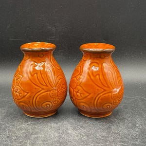 May include: Two orange ceramic vases with a glossy finish. Each vase has a rounded body and a narrow neck with a brown rim. The vases are decorated with a floral and leaf pattern.