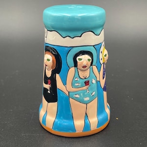 May include: A ceramic salt shaker with a light blue top and a cylindrical body. The body is painted with three figures in swimsuits, holding glasses of red liquid. The background is blue with white accents.
