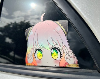 Può includere: Un adesivo colorato in stile anime di un personaggio con capelli rosa, orecchie da gatto e grandi occhi verdi, che sbuca da un finestrino dell'auto. L'adesivo ha un effetto olografico e fa il segno della pace con entrambe le mani.