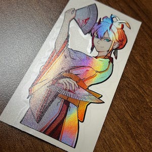 May include: A holographic sticker featuring a woman with blue and red hair holding a fan. She is wearing a white and rainbow kimono with a floral pattern.
