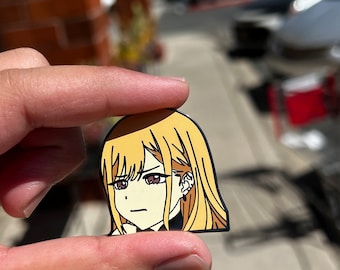 Può includere: Uno spillo in smalto dorato con un personaggio in stile anime con capelli biondi, occhi marroni e un'espressione seria. Il personaggio indossa una camicia bianca e nera con colletto.
