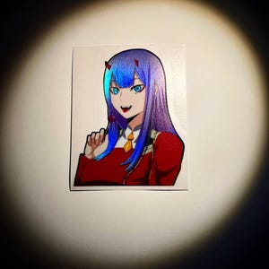 May include: A sticker featuring an anime character with blue and purple hair, red eyes, and a red and white outfit. The character is smiling and holding a lollipop.