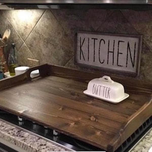 May include: A dark stained wooden stove top cover with handle cutouts. A white butter dish with the word "BUTTER" is on the cover. A sign that says "KITCHEN" is in the background.
