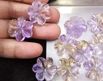 May include: Assortment of carved flower-shaped gemstones in shades of purple and yellow. The gemstones are arranged on a white surface.