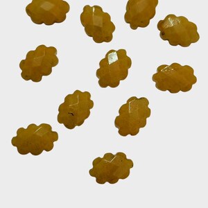 Pode incluir: Uma coleção de onze contas de pedras preciosas facetadas, amarelo-alaranjadas. Cada conta tem uma forma única, semelhante a uma nuvem, com múltiplas facetas. As contas estão espalhadas sobre um fundo branco, mostrando sua cor e corte.