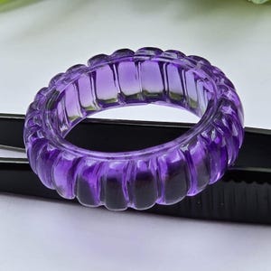 May include: A translucent purple ring carved from a single piece of amethyst. The band features a repeating vertical ribbed or fluted texture around its entire circumference, creating a scalloped edge profile with a polished, glossy finish.