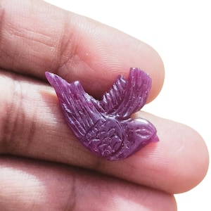 May include: A carved, deep magenta gemstone bird figurine. The bird is depicted in flight with detailed wing and feather carvings. The gemstone has a polished finish and is held in a hand.