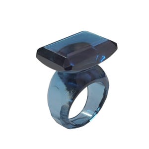 Puede incluir: Un anillo azul transparente con una gran gema rectangular. La banda del anillo es gruesa y facetada, y la gema es de un tono azul más oscuro. El anillo está sobre un fondo blanco.