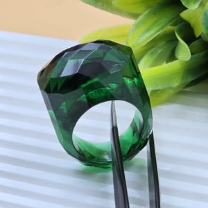 May include: A deep emerald green ring featuring a faceted, geometric design with a translucent, glass-like appearance. The ring is held by a pair of thin, dark metal tweezers, showcasing its angular cut and polished, reflective surface.
