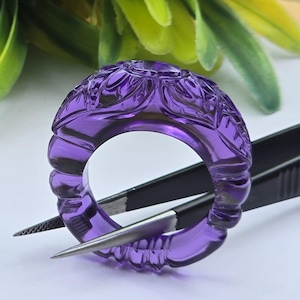 May include: A translucent purple amethyst ring featuring intricate floral carvings along the outer band. The ring is held by a pair of black metal tweezers, showcasing the deep violet hue and the detailed, textured surface of the gemstone material.
