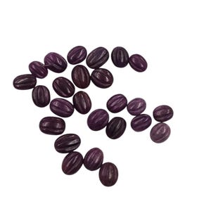 May include: A collection of oval-shaped, dark purple beads with carved, parallel lines. The beads are scattered across a white background, showcasing their textured surfaces and varying shades of purple.