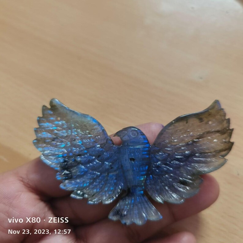Puede incluir: Una escultura de piedra de labradorita tallada de un p&aacute;jaro con las alas extendidas. La escultura presenta tonos iridiscentes azules, verdes y dorados. Las alas del p&aacute;jaro est&aacute;n detalladas con plumas talladas, y el cuerpo es de color azul oscuro.