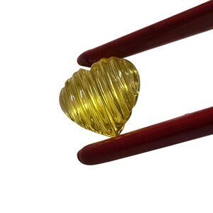 May include: A heart-shaped, yellow gemstone with a ridged surface is held by red tweezers. The gemstone has a translucent appearance and a bright, sunny color. The tweezers are a deep red.