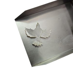 May include: A mother-of-pearl bird pendant with outstretched wings. The iridescent white pendant is set against a silver-colored metal tray. The bird's wings and tail feathers are intricately detailed.