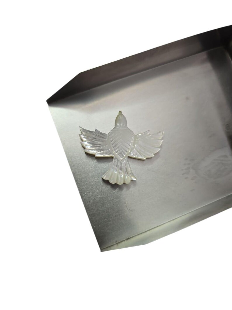 May include: A mother-of-pearl bird pendant with outstretched wings. The iridescent white pendant is set against a silver-colored metal tray. The bird's wings and tail feathers are intricately detailed.