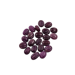 May include: A collection of oval-shaped, purple gemstones with carved, parallel lines. The stones vary in shades of purple, from light to dark, and are scattered against a white background. The gemstones appear to be polished.