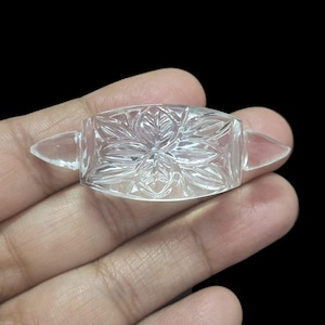 May include: A clear, rectangular crystal with a floral design carved into the center. The crystal has pointed ends and is held in a hand against a black background. The crystal is likely a decorative item or a component of jewelry.