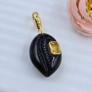May include: A black pendant with a carved design and a gold-coloured bail. A square, yellow gemstone is set in a gold-coloured bezel on the pendant. The pendant is likely made of a dark, polished material.