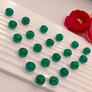 May include: A selection of small, round, emerald green beads with a ribbed, textured surface. The beads are scattered on a white, ridged surface, with red and pink flowers in the background. These beads are ideal for jewellery making.