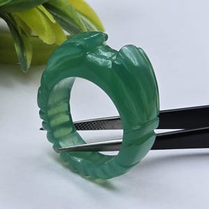 May include: A translucent green ring carved from a single piece of stone, featuring a series of vertical ridges and grooves along the outer band. The ring has a smooth, polished finish and is held by a pair of black metal tweezers.