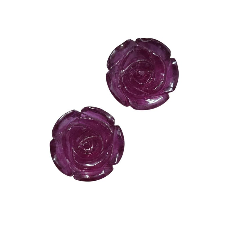 May include: Two carved rose-shaped gemstones in a deep purple hue. The detailed carvings showcase the petals and center of the roses. The gemstones are set against a plain white background.
