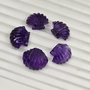 May include: Five carved amethyst gemstone seashell cabochons. The shells are a deep purple color and have detailed ridged textures. The cabochons are arranged in a circular pattern on a white surface with subtle lines.