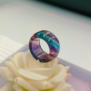 May include: A fluorite ring displaying bands of purple, blue, and green. The ring's colours are accentuated against a white backdrop. This gemstone ring showcases unique mineral patterns. A striking piece of jewellery.