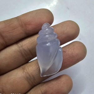 May include: A pale lavender chalcedony gemstone carved into the shape of a seashell. The seashell is detailed with ridges and swirls.