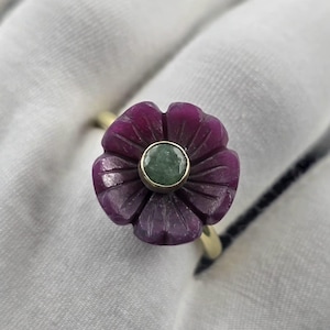 May include: A gold ring featuring a carved flower design with a deep magenta hue. The flower's center holds a small, round, green gemstone. The ring is set against a soft, white background.