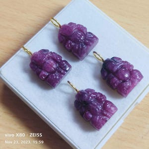 Pode incluir: Quatro pingentes de pedras preciosas roxas esculpidas com argolas douradas. Os pingentes têm um design floral e são exibidos em uma superfície branca. A imagem foi tirada com uma câmera vivo X80 Zeiss em 23 de novembro de 2023, às 15h59.