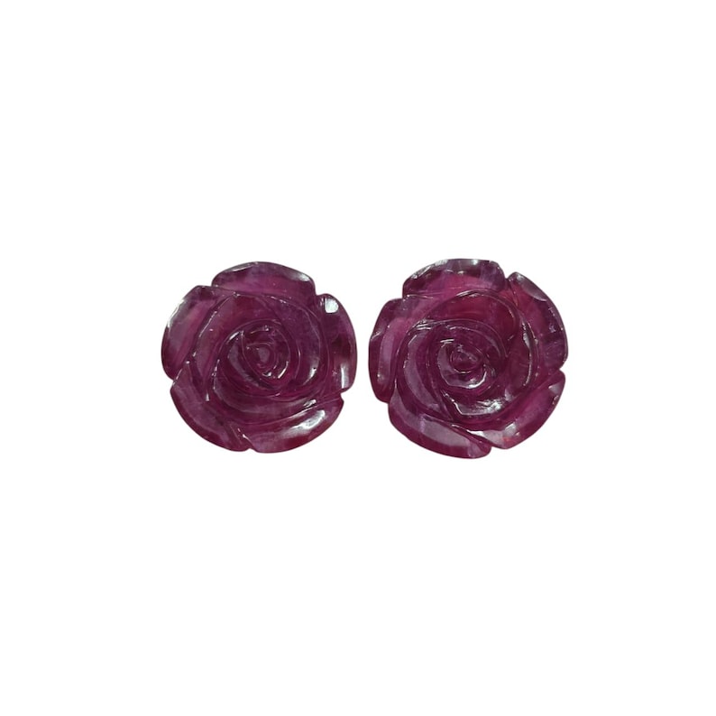 May include: Two carved rose-shaped gemstones in a deep magenta hue. The intricate details of the petals are visible, creating a realistic floral design. The gemstones are set against a plain white background.