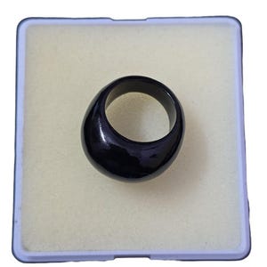 May include: A glossy black ring with a rounded, smooth design. The ring is set against a light beige background within a square, light blue-edged container. The ring's shape is a solid, curved band.