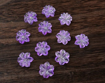 May include: A collection of twelve purple flower-shaped glass beads. The beads are arranged in a heart shape on a dark brown wooden surface.