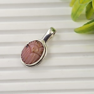 May include: A pink pendant with a carved design, set in a silver-colored metal frame. The pendant has an oval shape with a loop at the top for a chain. The carved design appears to be a floral or abstract motif.