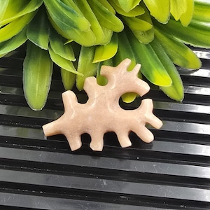 以下が含まれることがあります： 抽象的で枝分かれしたデザインの、薄茶色の彫刻された石の彫刻。彫刻は滑らかな質感で、緑の葉のある黒い背景に配置されています。