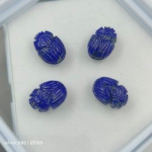 Puede incluir: Cuatro cuentas de piedras preciosas de lapislázuli talladas. Cada cuenta es de color azul oscuro con detalles tallados. Las cuentas están dispuestas en un cuadrado, sobre un fondo blanco.