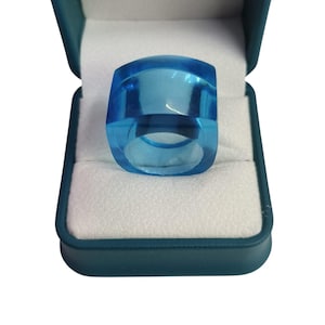 Puede incluir: Un anillo azul claro transparente con una parte superior rectangular y bordes redondeados, exhibido en una caja de anillo verde azulado con interior blanco. El anillo tiene un diseño geométrico moderno y está hecho de material transparente.